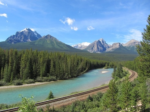 mountains with pine trees surrounding a small creek with a rail track along the water
