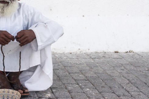 Old man sat cross legged on the ground with small basket in front of him. Man is holding prayer beads