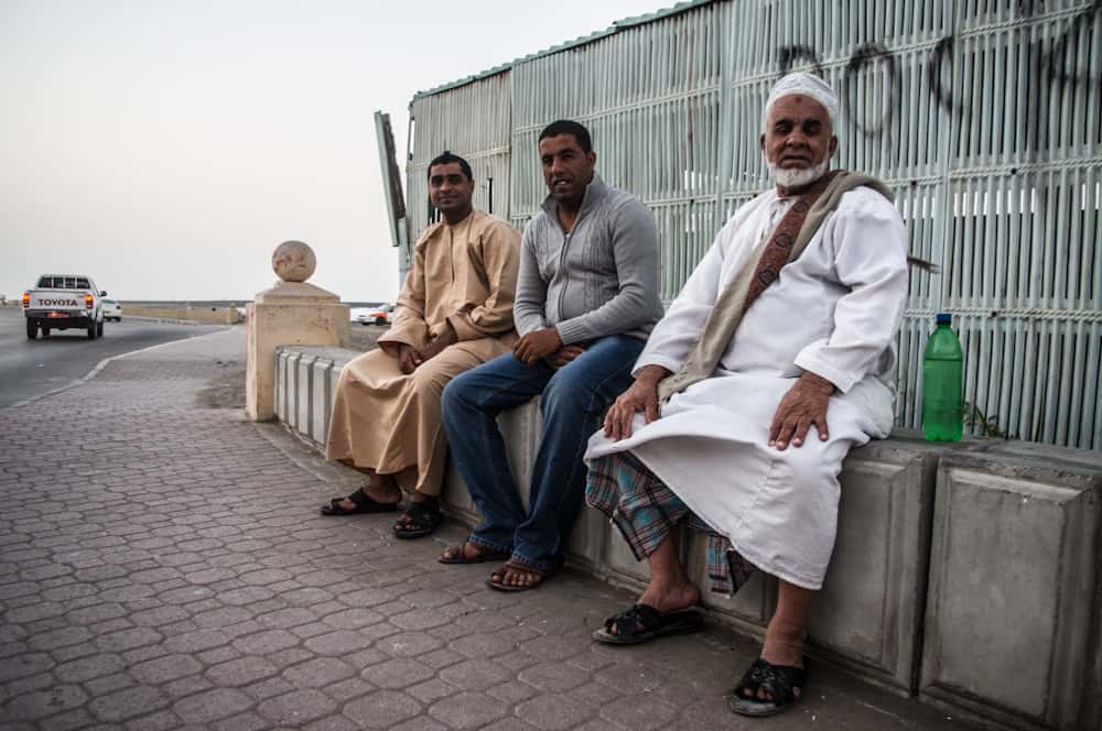 3 men sit on the side of the road with cars in the background