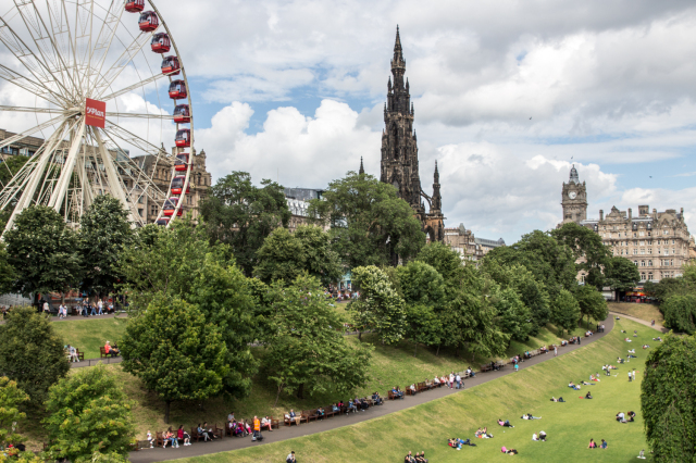 Picture of a park in Edinburgh with people out enjoying the sun with a castle and ferris wheel in the background