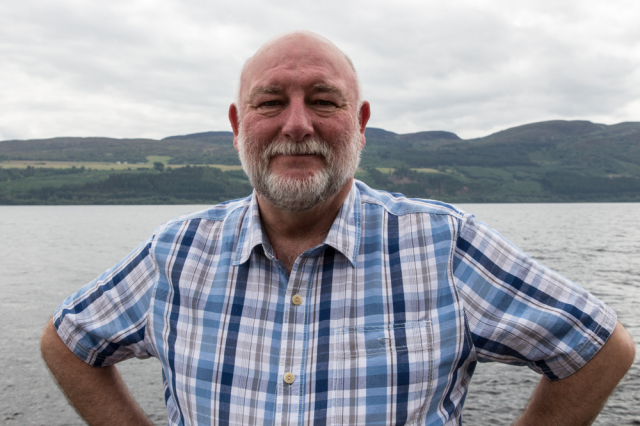 Alter Mann lächelt in einem blauen Hemd vor dem Loch Ness