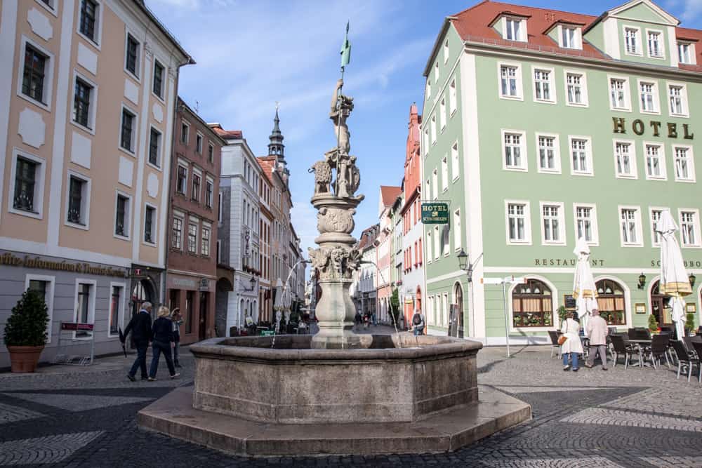 Town square with a large fountain in the middle, green hotel on the right and people walking down the street in Görlitz Obermarkt