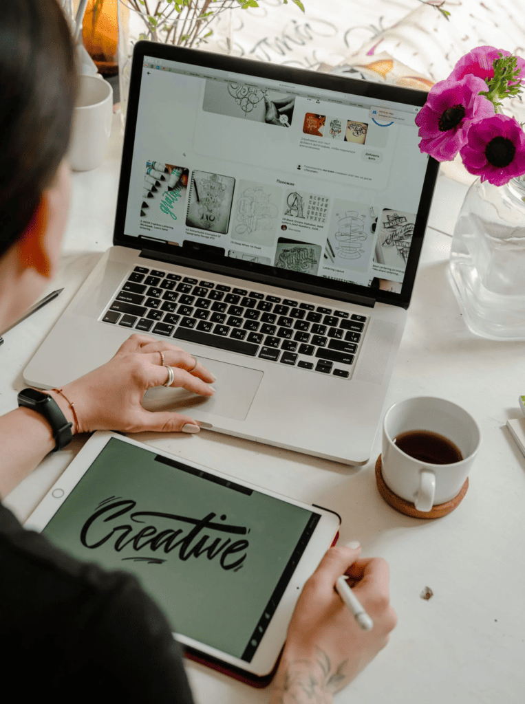 person using a laptop and tablet to create side hustle ideas with coffee and flowers