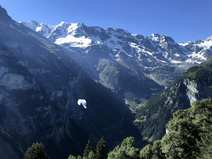 White paraglide shown paragliding in Switzerland over the Alps with snow covered mountains and greenery
