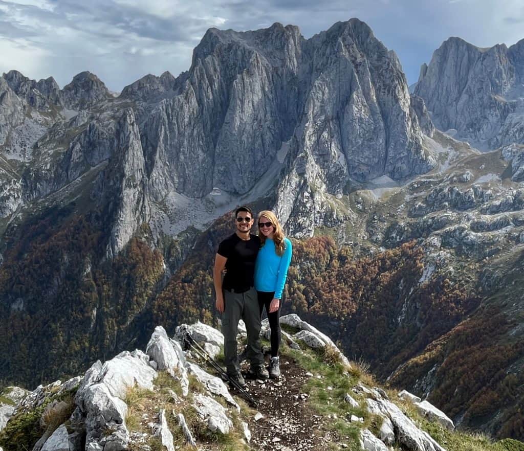 man and woman smiling on hike with large grey mountains behind them
