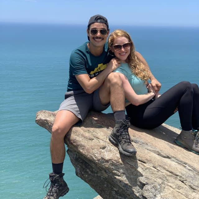 man and women smile after hike siting on a rock over looking blue ocean water