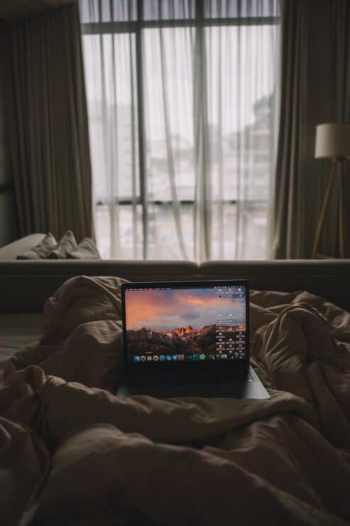 A Laptop in Bed with a window seeing a city landscape