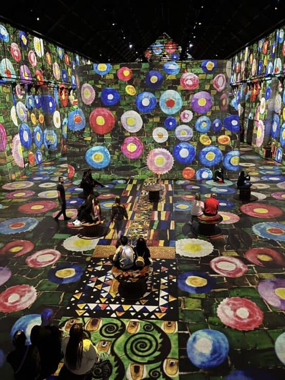 Colorful artistic circles fill the room projected on all walls celing and floor at fabrique des lumieres in amsterdam with kids and adults admiring the art
