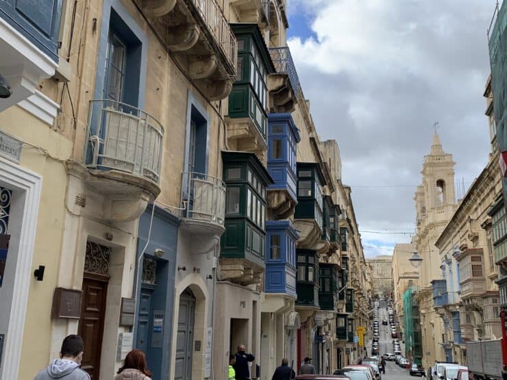 Colorful wooden balconies of green and blue line a busy street of cars and people in Malta