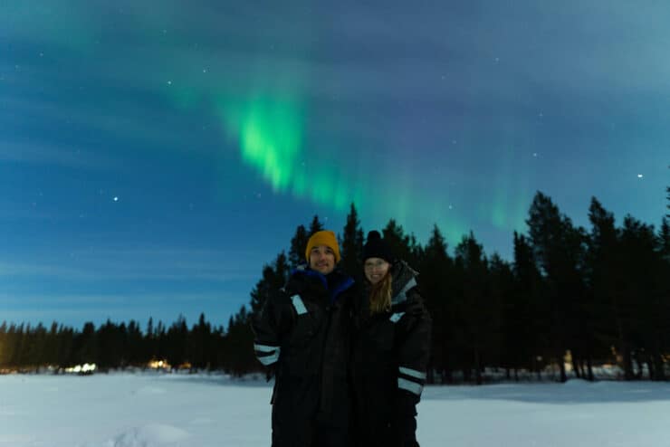 two people standing in the snow with Northern Lights across the background 