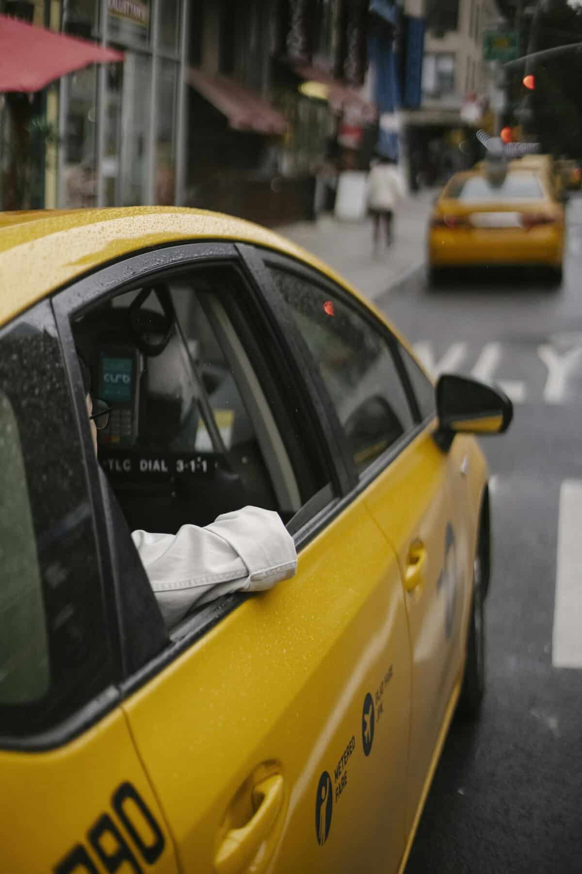 From above of unrecognizable person in white shirt sitting in modern yellow cab on asphalt roadway on urban background in town at daytime