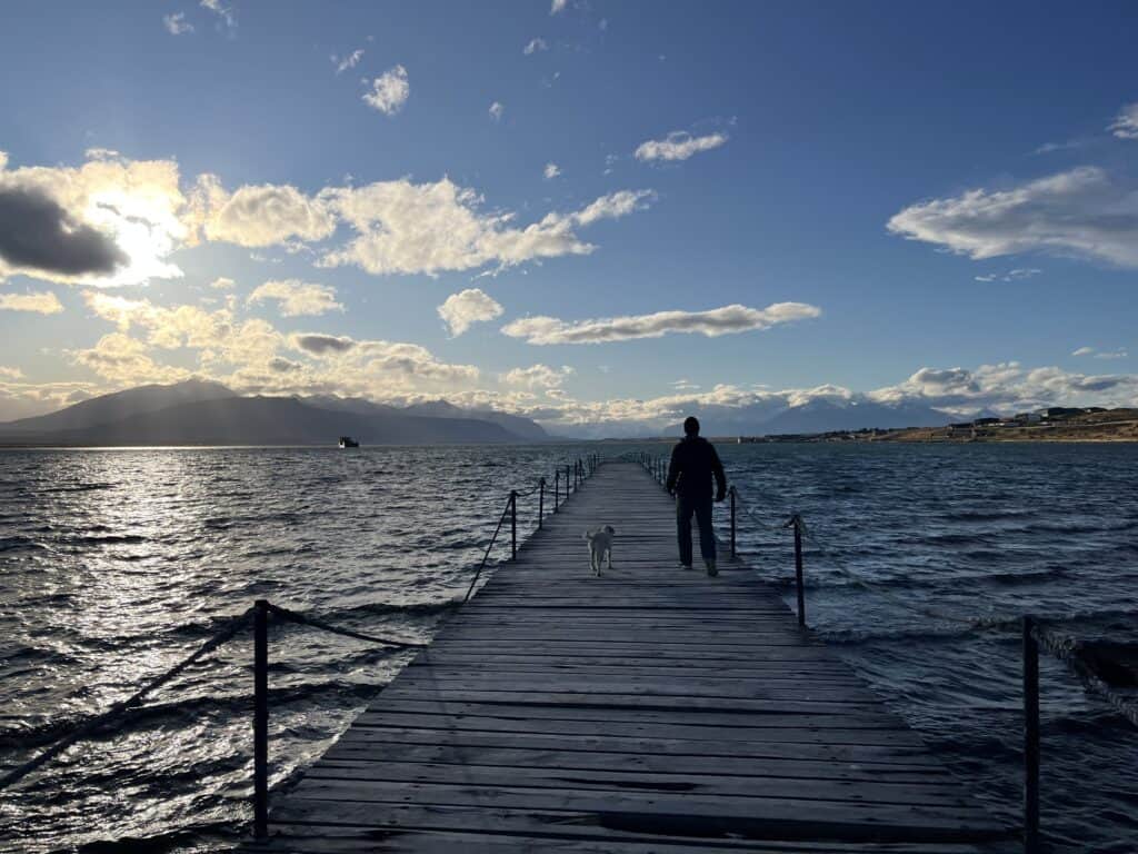 Man walks with dog on dock over water with mountains in the background