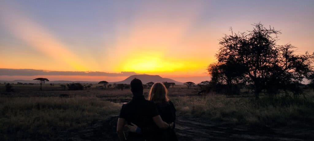 Couple sitting arm in arm watching an orange sunset over the African savanna, with acacia trees and a flat-topped hill in the distance, on the G Adventures Nairobi to Cape Town overland trip