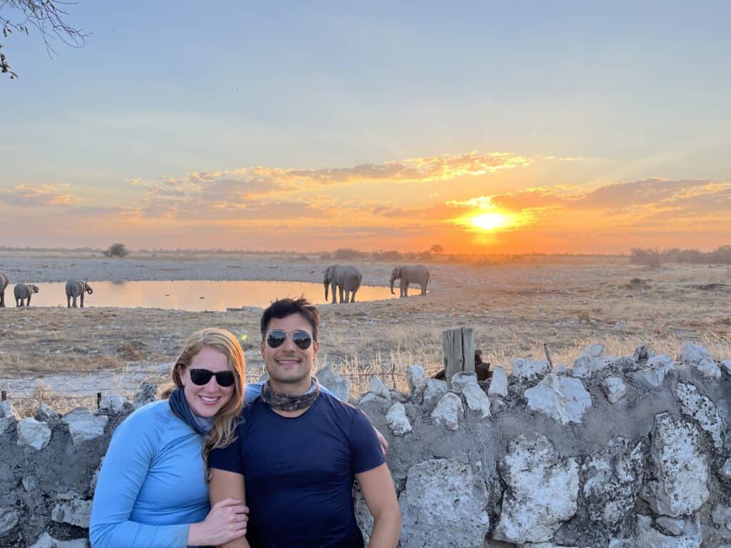 Couple in sunglasses smiling in front of a waterhole at sunset in Etosha National Park, Namibia, with a herd of elephants drinking behind them