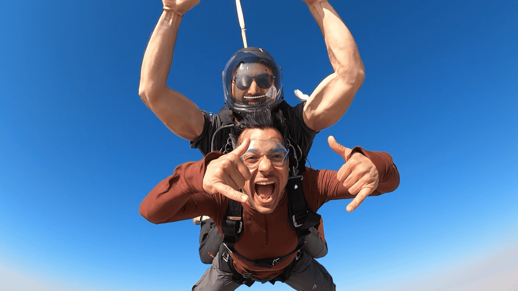 Tandem skydiver grinning and throwing hand signs in freefall against a clear blue sky, an optional activity on the Nairobi to Cape Town overland route